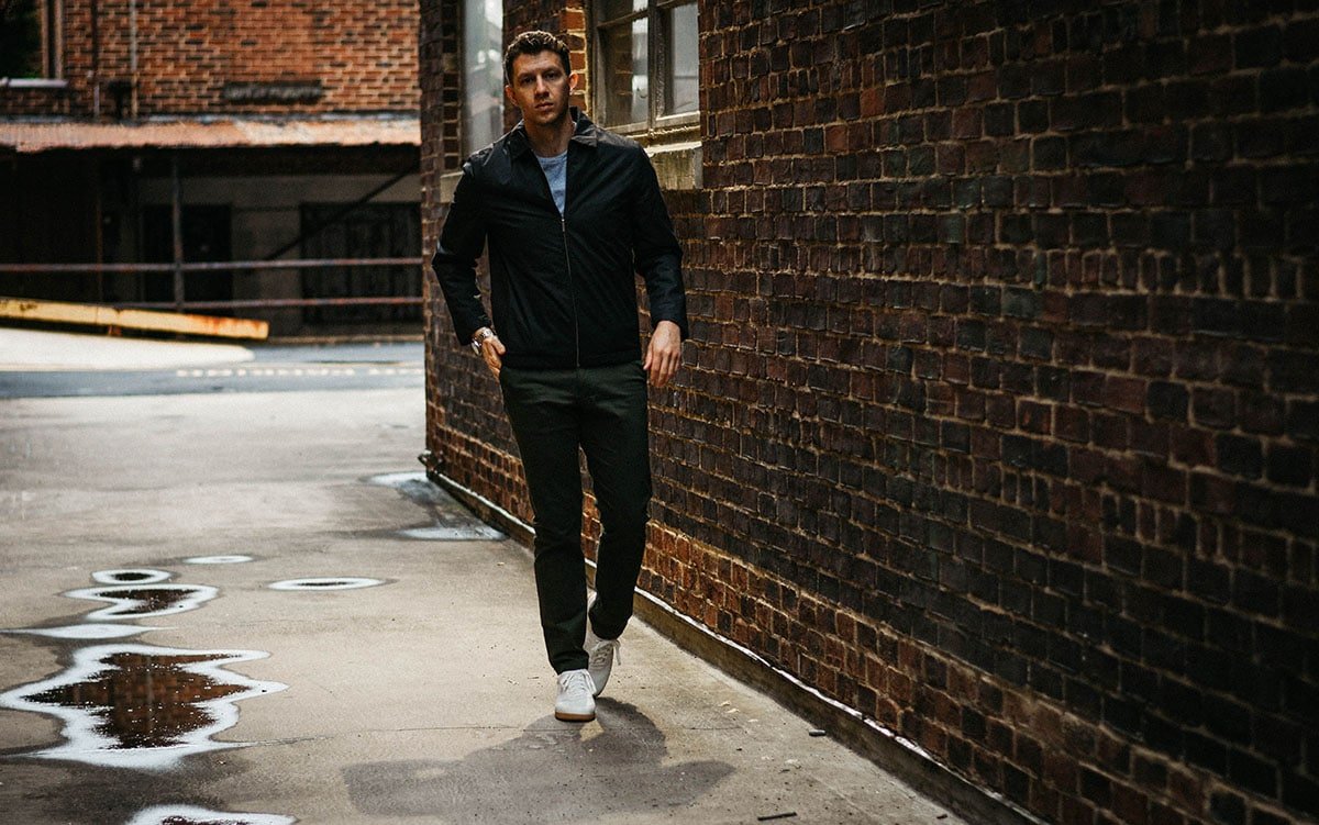 Asket Review model walking near brick wall in alley wearing black jacket olive chinos and white sneakers