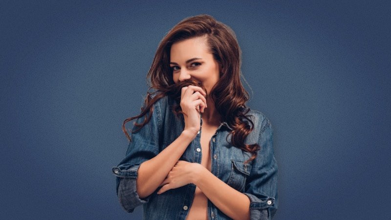 Attractive brunette woman smiling seductively at camera on blue background in jean jacket 1