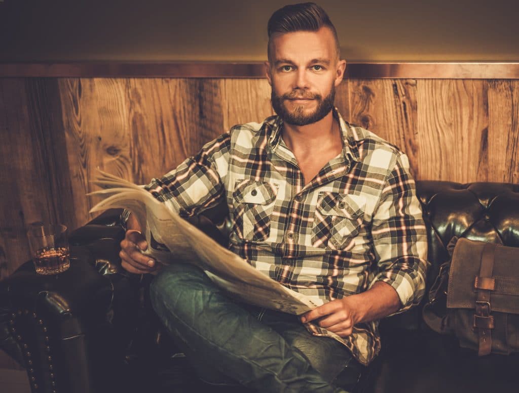 Middle-aged hipster reading newspaper on leather sofa in barber shop