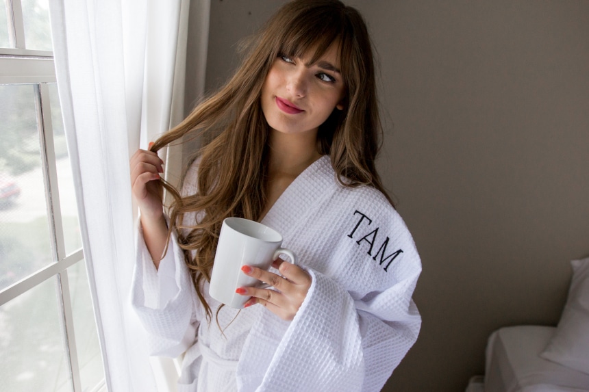 Female model wearing Luxor Linens Lakeview Signature Egyptian Cotton Spa Robe while holding a coffee and looking out the window and twirling her hair