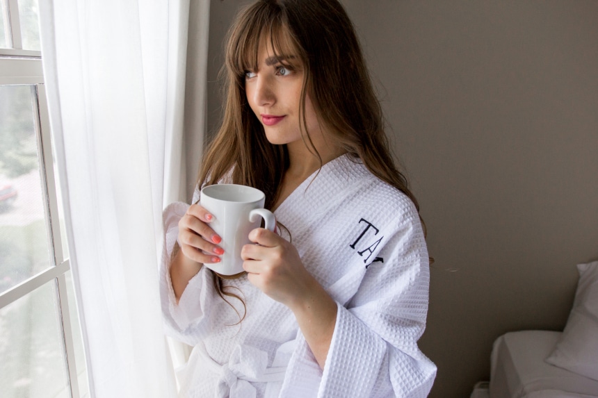 Female model wearing Luxor Linens Lakeview Signature Egyptian Cotton Spa Robe while holding a coffee and looking out the window