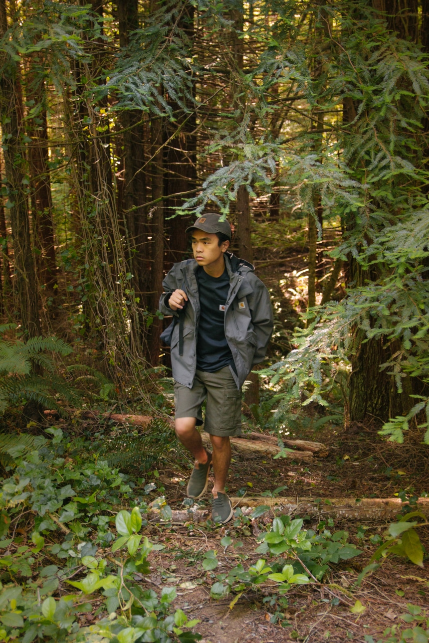 Man hiking through woods and carrying backpack wearing Carhartt Odessa Cap Rugged Flex Rigby Cargo Shorts Force Delmont Tee and Shoreline Jacket a