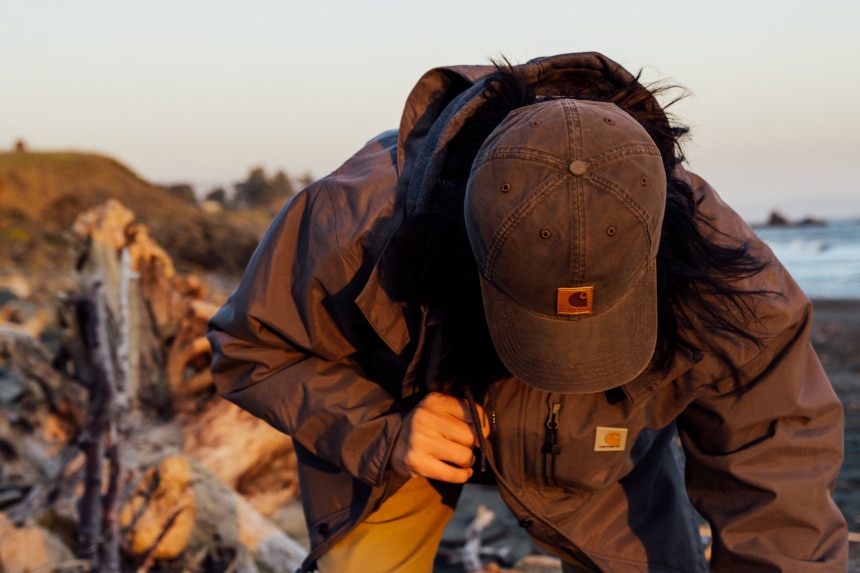 Man looking at driftwood on a beach while wearing Carhartt Shoreline Jacket Odessa Cap and Rugged Flex Rigby Dungaree