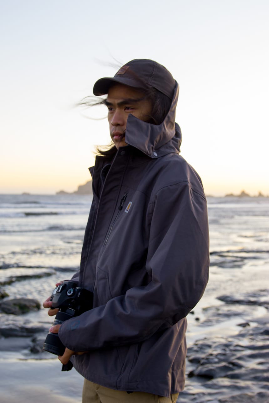 Man on beach next to ocean holding camera in the wind while wearing Carhartt Shoreline Jacket and Rugged Flex Dungaree
