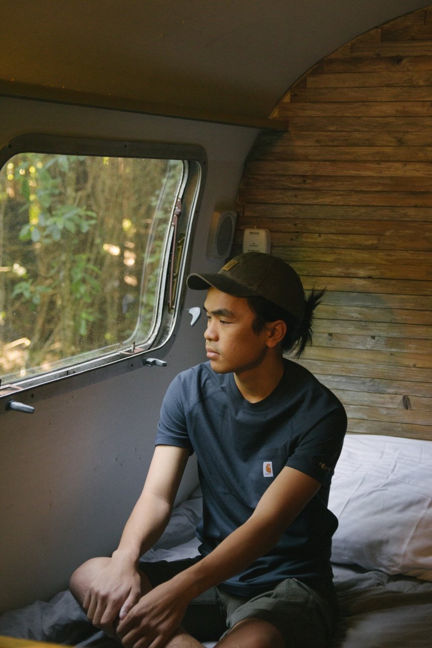 Man sitting in Airstream camper on bed looking out the window wearing Carhartt Odessa Cap Force Delmont Tee and Rugged Flex Cargo Shorts a