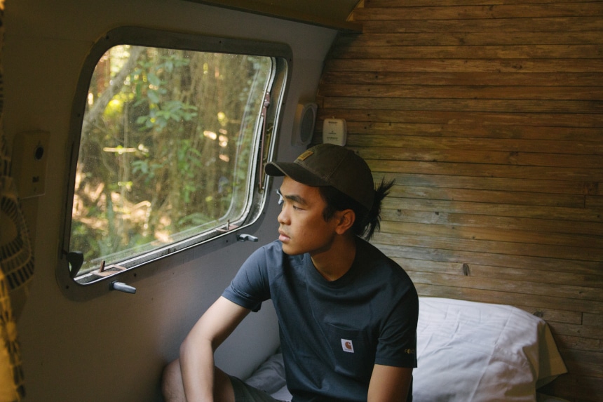 Man sitting in Airstream camper on bed looking out the window wearing Carhartt Odessa Cap Force Delmont Tee and Rugged Flex Cargo Shorts b