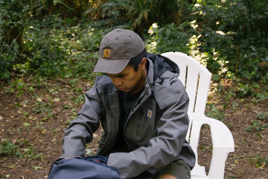 Man sitting on chair looking through backpack while wearing Carhartt Shoreline Jacket Odessa Cap and Rugged Flex Rigby Dungaree
