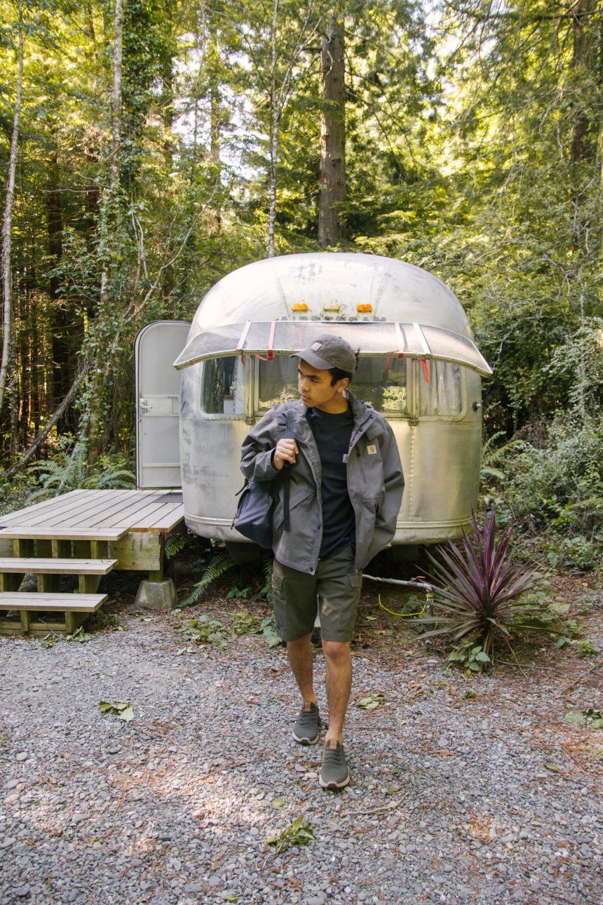 Man walking away from Airstream camper in the woods wearing backpack and Carhartt Odessa Cap Shoreline Jacket Rugged Flex Rigby Cargo shorts and Force Delmont Tee