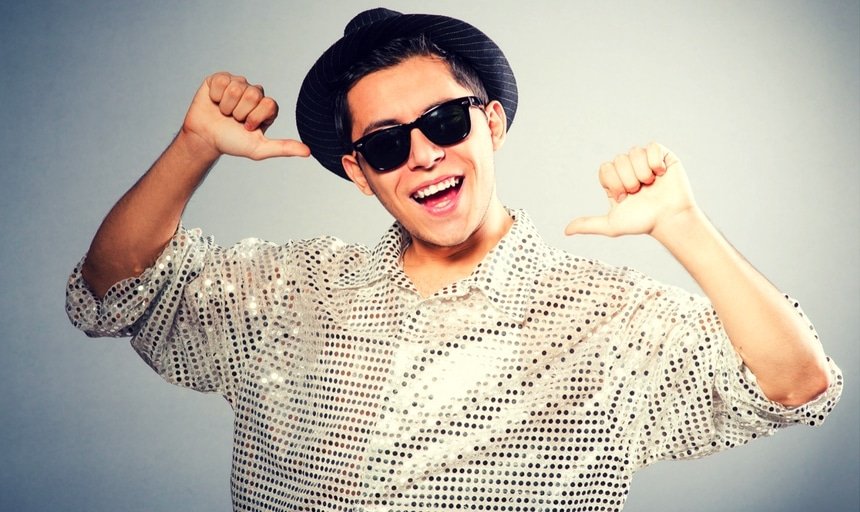 Man with shiny shirt and fedora pointing to himself and smiling