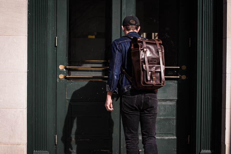 model entering large green doors wearing rolltop leather backpack moral code