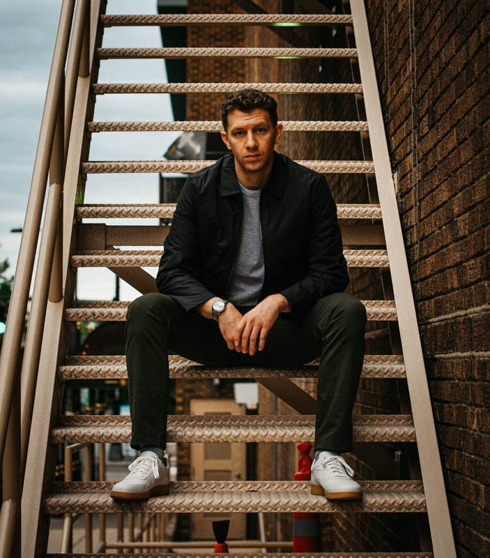 model sitting on steel staircase wearing olive chinos zip jacket and white sneakers
