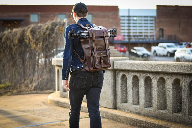 Model wearing leather rolltop backpack on bridge with hat