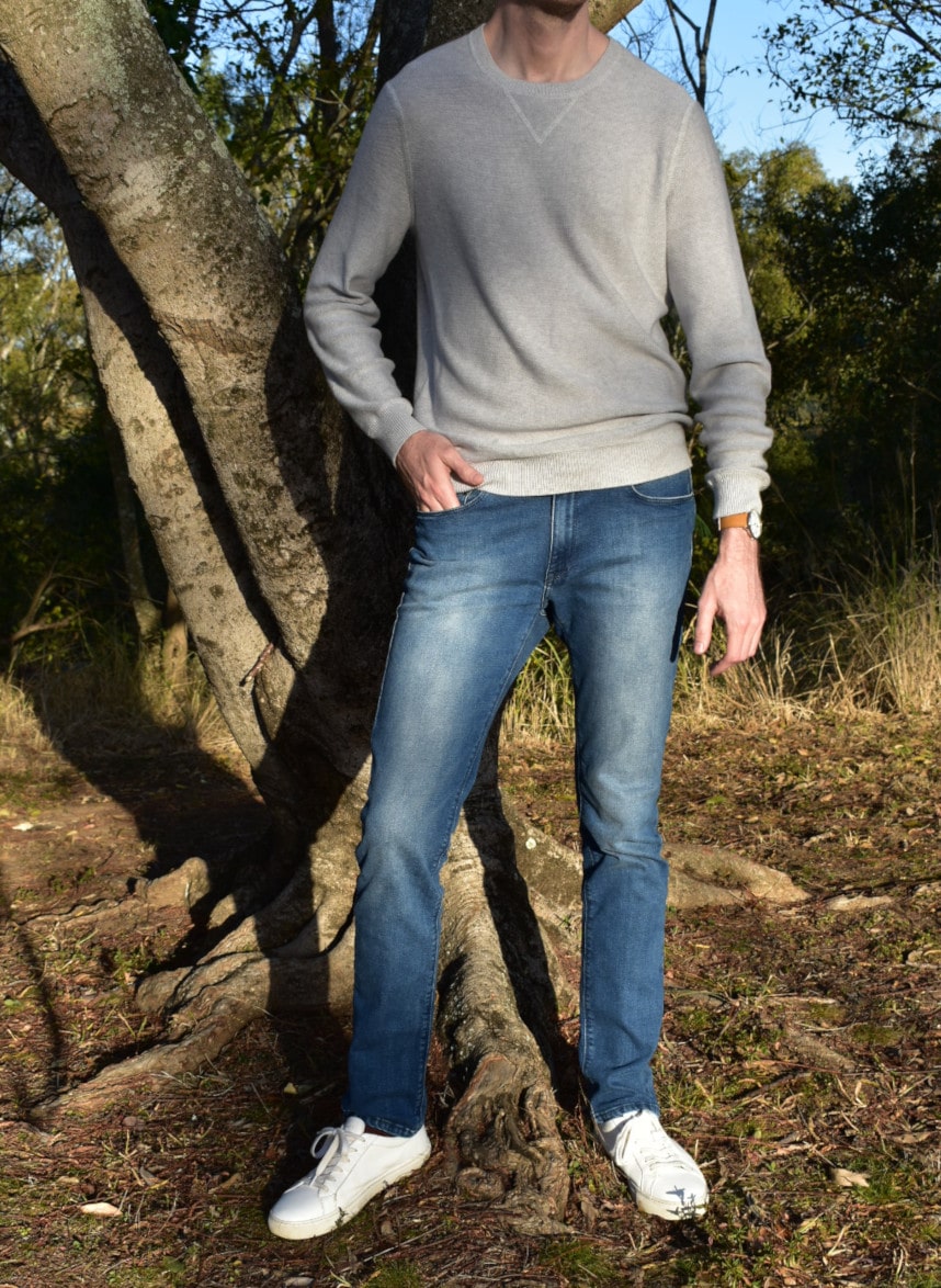 Model Wearing Mott & Bow Slim Laight Jeans with Liam Sweater Standing Up and Standing Next to Tree With Hand in Pocket and with White Sneakers