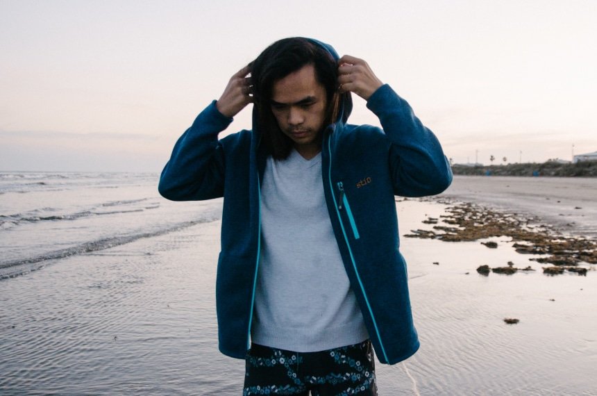 Model Wearing Stio Wilcox Fleece Hoodie and CFS Board Shorts While Holding Hood at the Beach